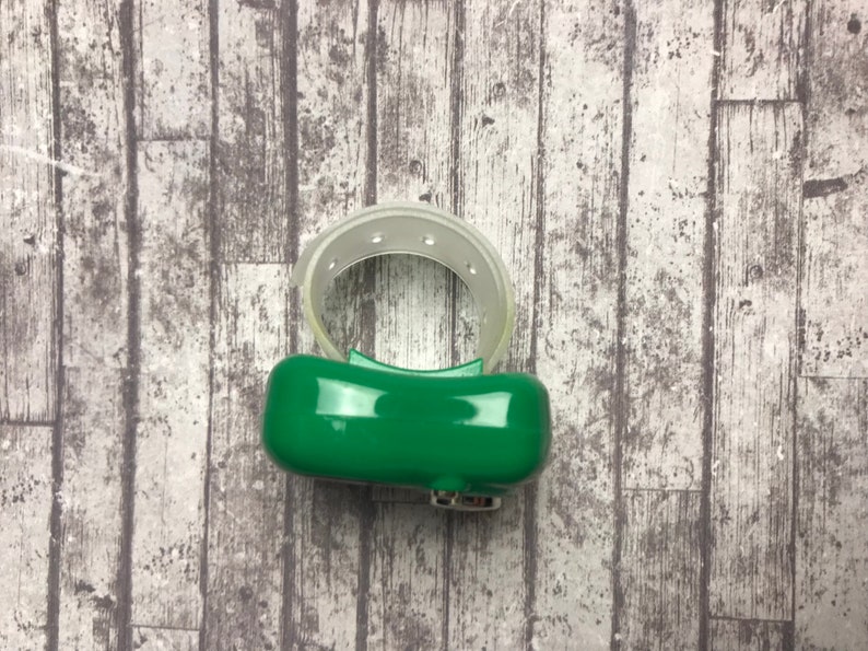 May include: A green and clear plastic ring with a small button on the side. The ring is designed to be worn on the finger.