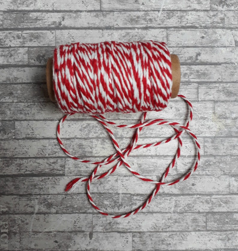 May include: A roll of red and white baker's twine with a loose end. The twine is on a white wood background.