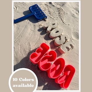 Może przedstawiać: Scena na plaży z niebieskimi grabiami i słowem "rose" uformowanym w piasku. Jasnoróżowe foremki do piasku w kształcie cyfr wymawiają "1309". Białe koło z tekstem "10 kolorów dostępnych" znajduje się w lewym dolnym rogu.