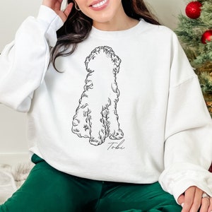 May include: White sweatshirt featuring a black and white illustration of a dog. The dog illustration has a fluffy appearance. The name "Tobi" is written in cursive below the dog. The person in the image is wearing green pants.