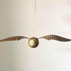 May include: A golden Snitch, a spherical object with two wings, hangs suspended against a white background. The wings are detailed with rectangular segments, and the ball has a textured surface.