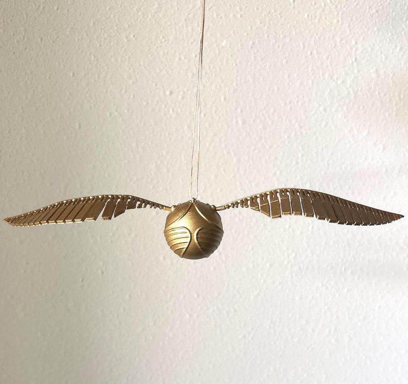 May include: A golden Snitch, a spherical object with two wings, hangs suspended against a white background. The wings are detailed with rectangular segments, and the ball has a textured surface.
