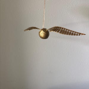May include: A golden Snitch ornament from the Harry Potter series, suspended by a thin string. The ball is round with textured details, and it has two sets of wings extending outwards. The wings are made of individual, rectangular pieces.