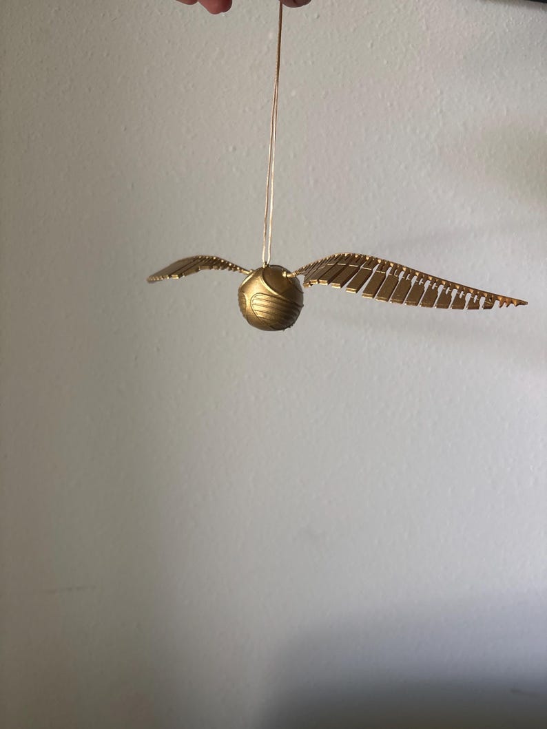 May include: A golden Snitch ornament from the Harry Potter series, suspended by a thin string. The ball is round with textured details, and it has two sets of wings extending outwards. The wings are made of individual, rectangular pieces.