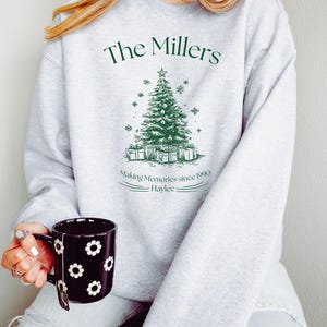 May include: A light gray sweatshirt with a green Christmas tree design and the text "The Millers". Below the tree are wrapped gifts, and the text "Making Memories since 1990 - Haylee". A black mug with white flower designs is held in the hand.