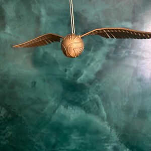 May include: A golden Snitch, a fictional flying ball from the Harry Potter series, hangs suspended by a thin string. The ball has detailed wings and a textured surface, set against a teal and green background.