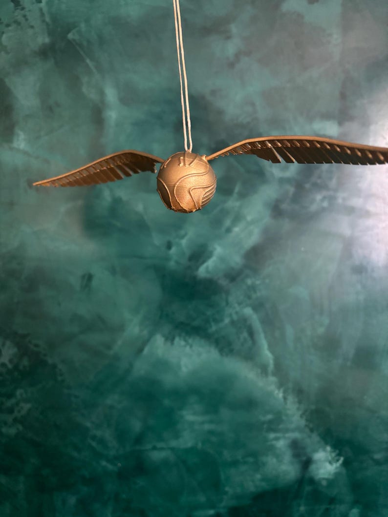 May include: A golden Snitch, a fictional flying ball from the Harry Potter series, hangs suspended by a thin string. The ball has detailed wings and a textured surface, set against a teal and green background.