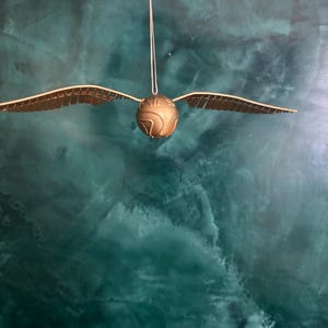May include: A golden Snitch ornament with outstretched wings, suspended by a string. The spherical body has etched details, and the wings are detailed with a series of small lines. The background is a mottled teal color.