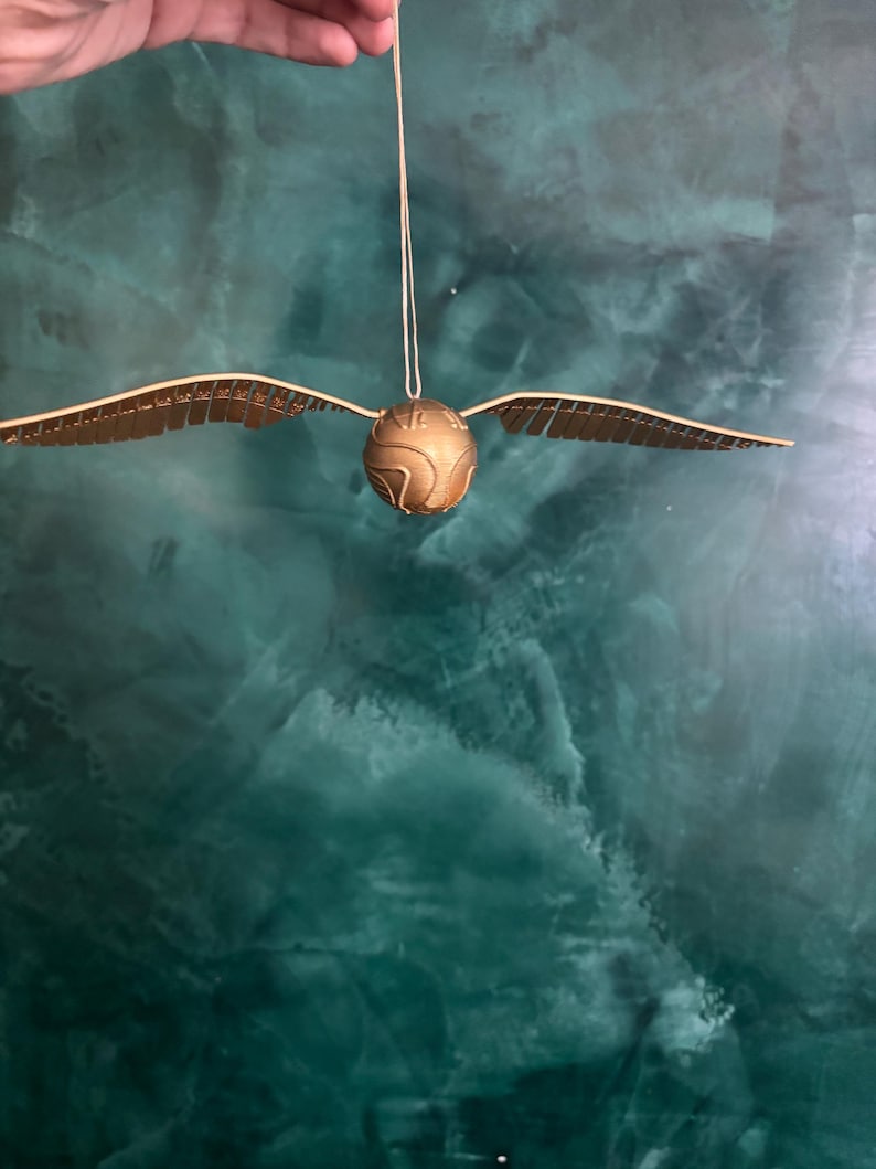 May include: A golden Snitch ornament with outstretched wings, suspended by a string. The spherical body has etched details, and the wings are detailed with a series of small lines. The background is a mottled teal color.
