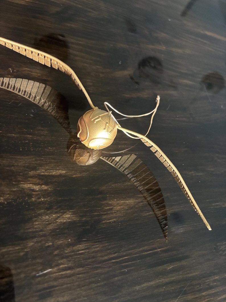 May include: A golden Snitch, a spherical object with wings, from the Harry Potter series. The wings are detailed with a textured pattern, and the sphere has engraved lines. A thin, white wire extends from the sphere.