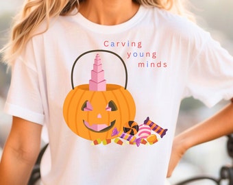 May include: White t-shirt with a Halloween design. The graphic shows an orange pumpkin bucket filled with pink blocks and surrounded by sweets. The text "Carving young minds" is printed above the bucket in red and blue.