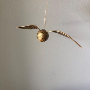 May include: A golden Snitch ornament with outstretched wings, suspended by a thin string. The ball is detailed with etched lines and the wings have a feathered texture. This decorative item is inspired by the wizarding world.
