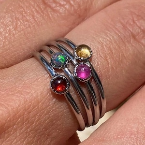 May include: Five silver rings with colorful gemstones. The rings are stacked on a finger. The gemstones are red, pink, yellow, and iridescent.