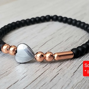 Peut inclure: Bracelet en perles noires avec un pendentif en forme de cœur argenté et des accents en or rose. Le bracelet porte une étiquette "Solazur" avec le texte "I ♥ U".