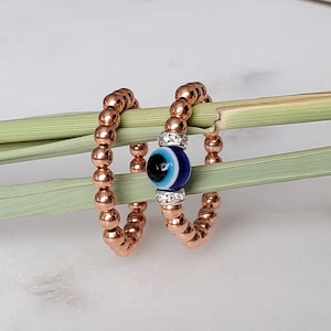 Puede incluir: Dos anillos de cuentas de oro rosa con un dije de ojo turco azul. El dije tiene un borde blanco y detalles plateados.