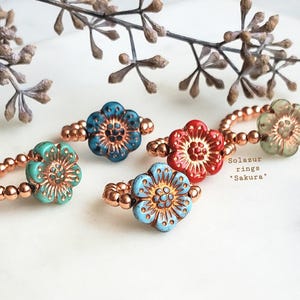 May include: Five copper rings with colorful flower-shaped charms. The rings are made of small copper beads and the charms are in shades of blue, green, red, and light blue.