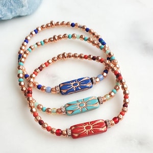 May include: Three beaded bracelets featuring metallic copper-toned beads interspersed with small blue, turquoise, and red beads. Each bracelet centres on a rectangular ceramic bead in blue, turquoise, or red, each displaying a gold sunburst design.