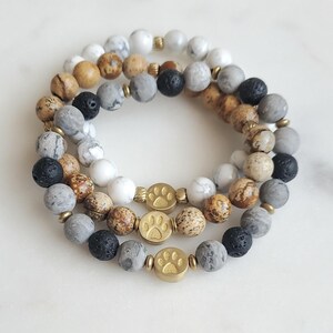 Peut inclure: Trois bracelets superposés avec des perles brunes, grises, blanches et noires. Chaque bracelet a un charme en forme de patte d'or.