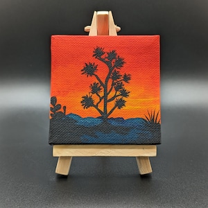 May include: A small canvas painting on an easel depicts a silhouette of a Joshua tree against a sunset background. The sky is painted in shades of orange, yellow, and red, while the tree is black. The painting is set against a dark background.