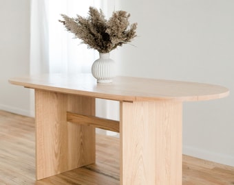 May include: A light wood oval table with a simple design. The table has a single, horizontal support beam connecting the two legs. The table is on a light wood floor.