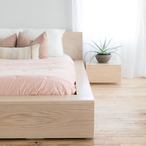 May include: A light pink bedspread with white pillows on a wooden platform bed frame. A small wooden nightstand with a potted plant sits next to the bed.