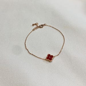 May include: A delicate rose gold bracelet featuring a four-leaf clover charm with a vibrant red center. The bracelet has a fine chain and an adjustable clasp. The clover charm is approximately 0.5 inches in diameter.