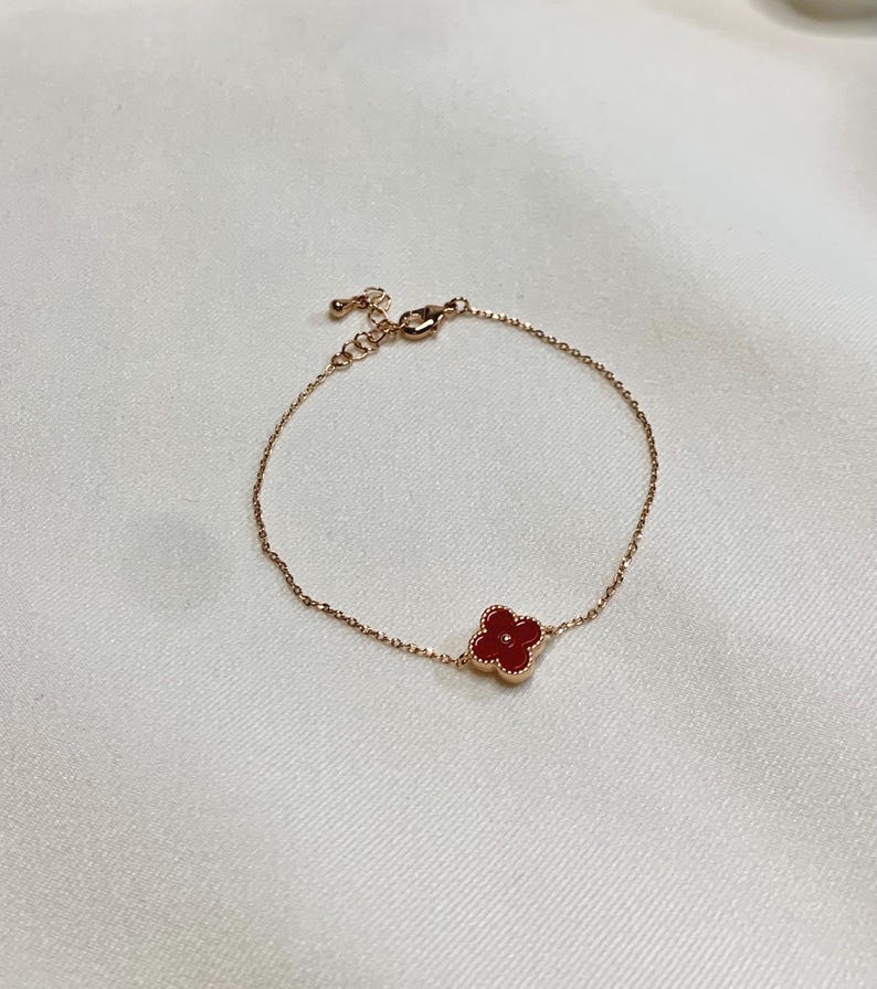May include: A delicate rose gold bracelet featuring a four-leaf clover charm with a vibrant red center. The bracelet has a fine chain and an adjustable clasp. The clover charm is approximately 0.5 inches in diameter.