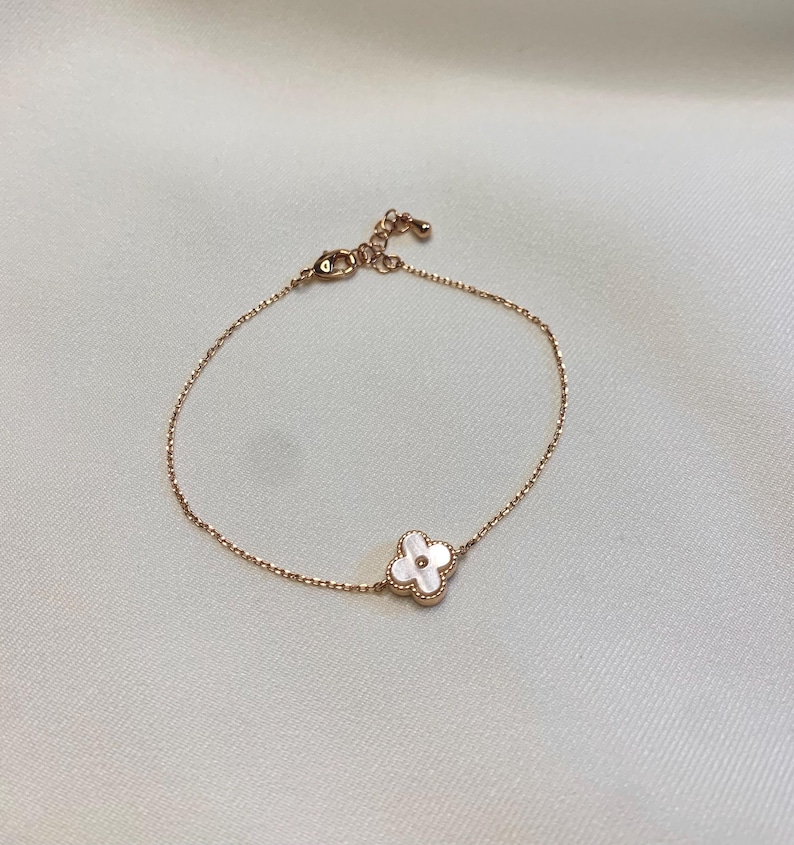 May include: A delicate gold-toned bracelet featuring a four-leaf clover charm with a white mother-of-pearl inlay. The bracelet has a fine chain and an adjustable clasp. The clover charm is centered on the bracelet.