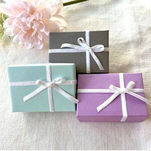 May include: Three small gift boxes in pastel colors: mint green, lavender, and gray. Each box is tied with a white satin ribbon and bow. A pink flower is in the background.