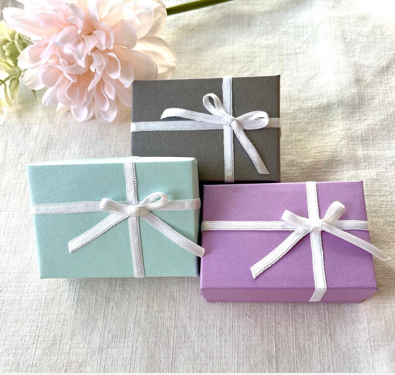 May include: Three small gift boxes in pastel colors: mint green, lavender, and gray. Each box is tied with a white satin ribbon and bow. A pink flower is in the background.