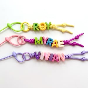 May include: Three colorful beaded name keychains. The names "ROY", "MYRAE", and "VIVIAN" are spelled out with multi-colored letter beads. Each keychain has a different color scheme, including green, pink, and purple.