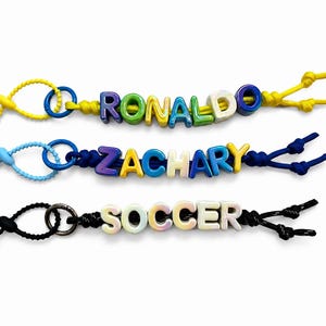 May include: Three personalized zipper pulls featuring colorful letter beads spelling RONALDO, ZACHARY, and SOCCER. Each pull is attached to a cord with a circular metal ring and a plastic toggle, available in yellow, blue, and black cord colors.