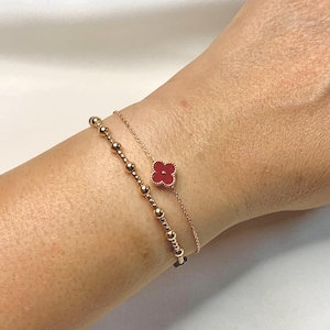 May include: Two delicate gold bracelets adorn a wrist. One features a series of small, round beads. The other has a red clover-shaped charm. The bracelets are set against a neutral background, highlighting the jewelry's design.