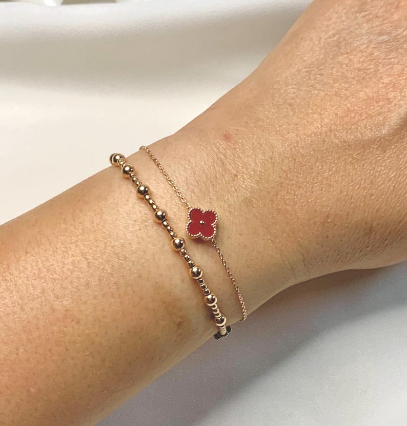 May include: Two delicate gold bracelets adorn a wrist. One features a series of small, round beads. The other has a red clover-shaped charm. The bracelets are set against a neutral background, highlighting the jewelry's design.