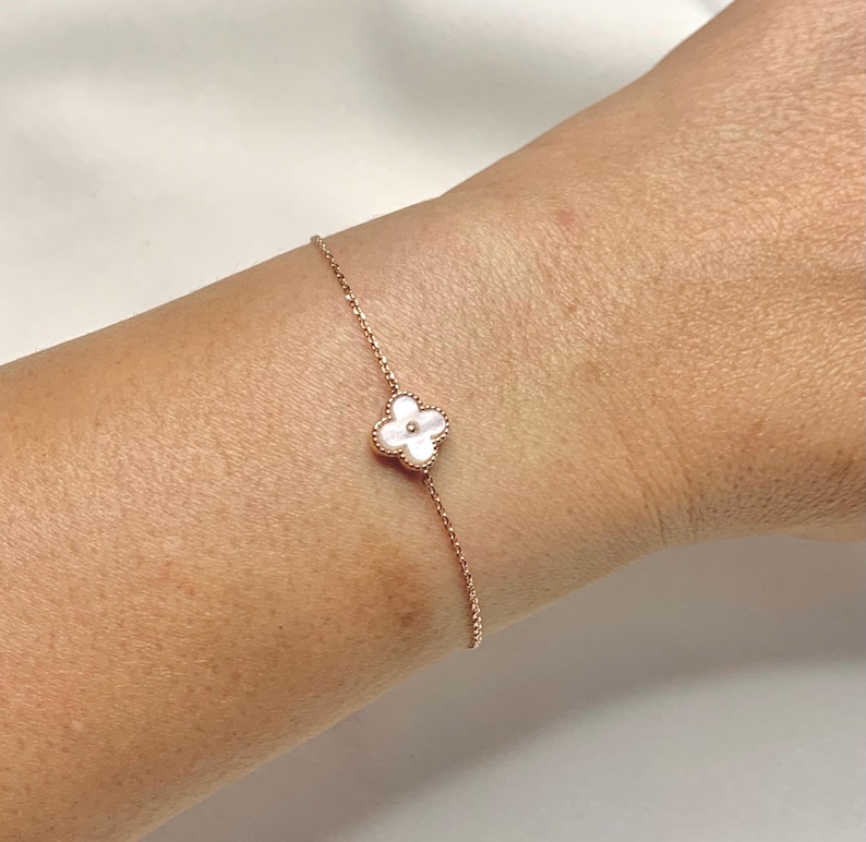 May include: Rose gold bracelet featuring a four-leaf clover charm with a white mother-of-pearl center. The delicate chain bracelet is displayed on a wrist against a white background. A piece of jewelry.
