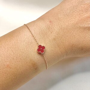 May include: A delicate gold-tone bracelet featuring a four-leaf clover charm with a vibrant red center. The clover design is set against a gold chain, creating a simple yet elegant accessory. The bracelet is shown on a wrist.