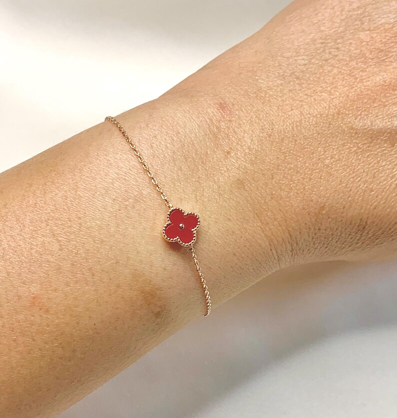 May include: A delicate gold-tone bracelet featuring a four-leaf clover charm with a vibrant red center. The clover design is set against a gold chain, creating a simple yet elegant accessory. The bracelet is shown on a wrist.