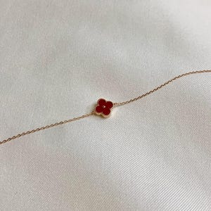 May include: A delicate gold-toned bracelet featuring a four-leaf clover charm with a vibrant red center. The clover is centered on the chain, and the bracelet is set against a soft, white background.