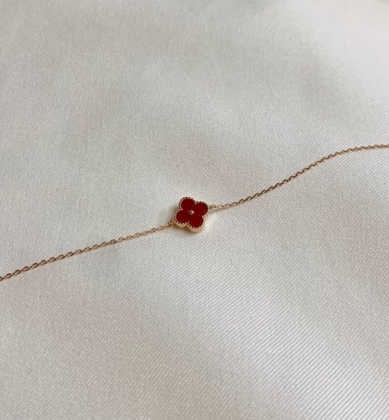 May include: A delicate gold-toned bracelet featuring a four-leaf clover charm with a vibrant red center. The clover is centered on the chain, and the bracelet is set against a soft, white background.