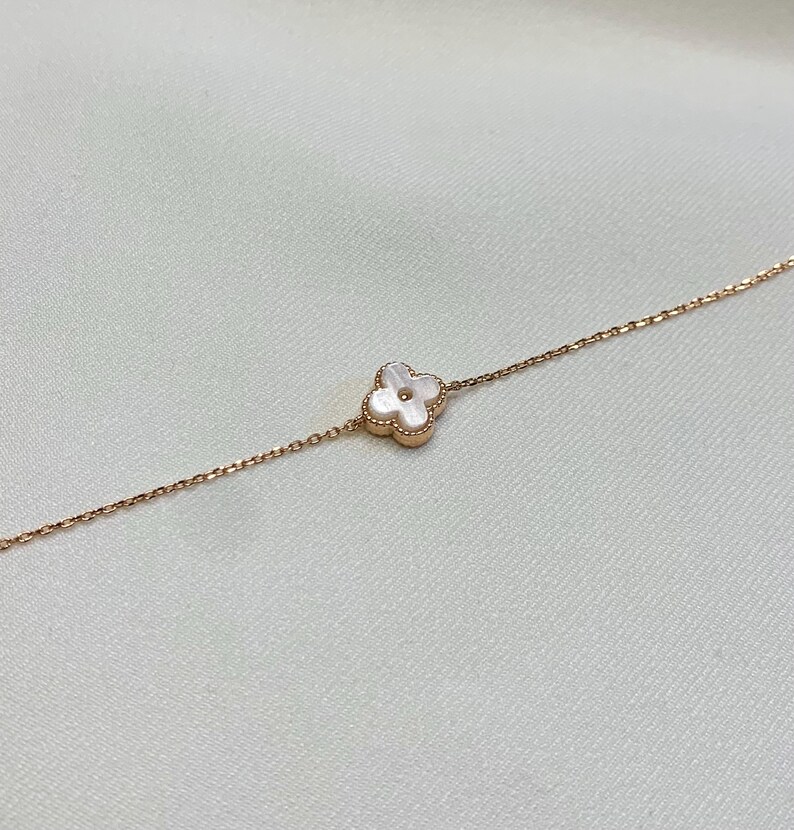 May include: A delicate rose gold bracelet featuring a four-leaf clover charm with a white mother-of-pearl center. The clover is set in a rose gold frame. The bracelet is displayed on a white background.