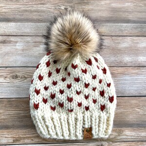 May include: Cream-colored knit beanie with a pattern of dark red hearts. The hat features a large, fluffy, faux fur pom-pom in shades of brown and tan. A small, rectangular tag with text is sewn on the brim.