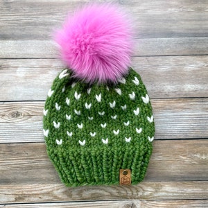 May include: A green knit beanie with white heart-shaped patterns and a large pink faux fur pom-pom. The hat has a small leather tag with a logo. This winter hat is a cozy accessory.