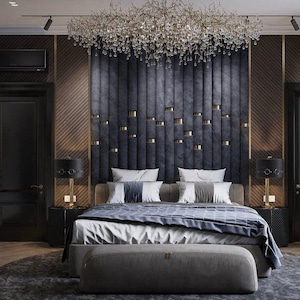 May include: A luxurious bedroom with a large bed featuring a gray headboard with gold accents. The bed has a white and gray patterned duvet and a gray footboard. The room has a large crystal chandelier and two bedside lamps with black shades.