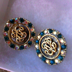 May include: A pair of gold-toned earrings with a circular design. Each earring features a dark grey centre with a gold monogram and is surrounded by a ring of dark green and iridescent rhinestones.