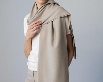 May include: A solid beige rectangular scarf with a smooth texture and delicate fringe detailing along the bottom edge. The scarf is draped over the shoulders and hangs down the front, showcasing a consistent neutral color and clean finish.