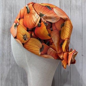 May include: A fabric head covering featuring a repeating pattern of orange pumpkins. The head covering is draped over a mannequin head, showcasing the fall-themed design. The pumpkins vary in size and are in shades of orange and yellow.