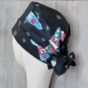 May include: A black scrub cap with a festive Christmas stocking pattern in red, blue, and green, accented with white snowflakes. The cap is tied in a bow at the back and is displayed on a mannequin head.