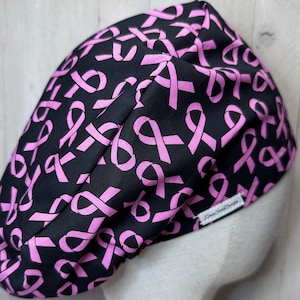 May include: A black surgical scrub cap featuring a repeating pattern of pink awareness ribbons. The cap is displayed on a mannequin head. The fabric has a small tag with text on the side. The cap is designed for medical professionals.