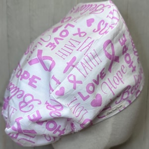 May include: White fabric cap featuring pink ribbons, hearts, and the words "Love, Faith, Hope, Believe". The cap's design includes a pattern of pink ribbons and words.