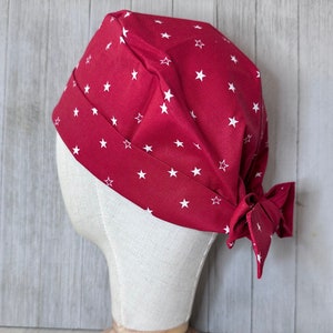 May include: Red fabric scrub hat with a white star pattern. The hat is tied at the back with a bow.